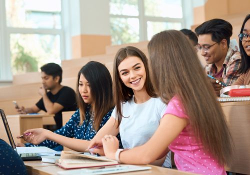Smiling students talking during lesson. Smiling students talking during lesson. Sitting in modern university classroom with wooden desks on groupmates background. Guys reading, studuing, writing. Boys and girls wearing casual clothes.